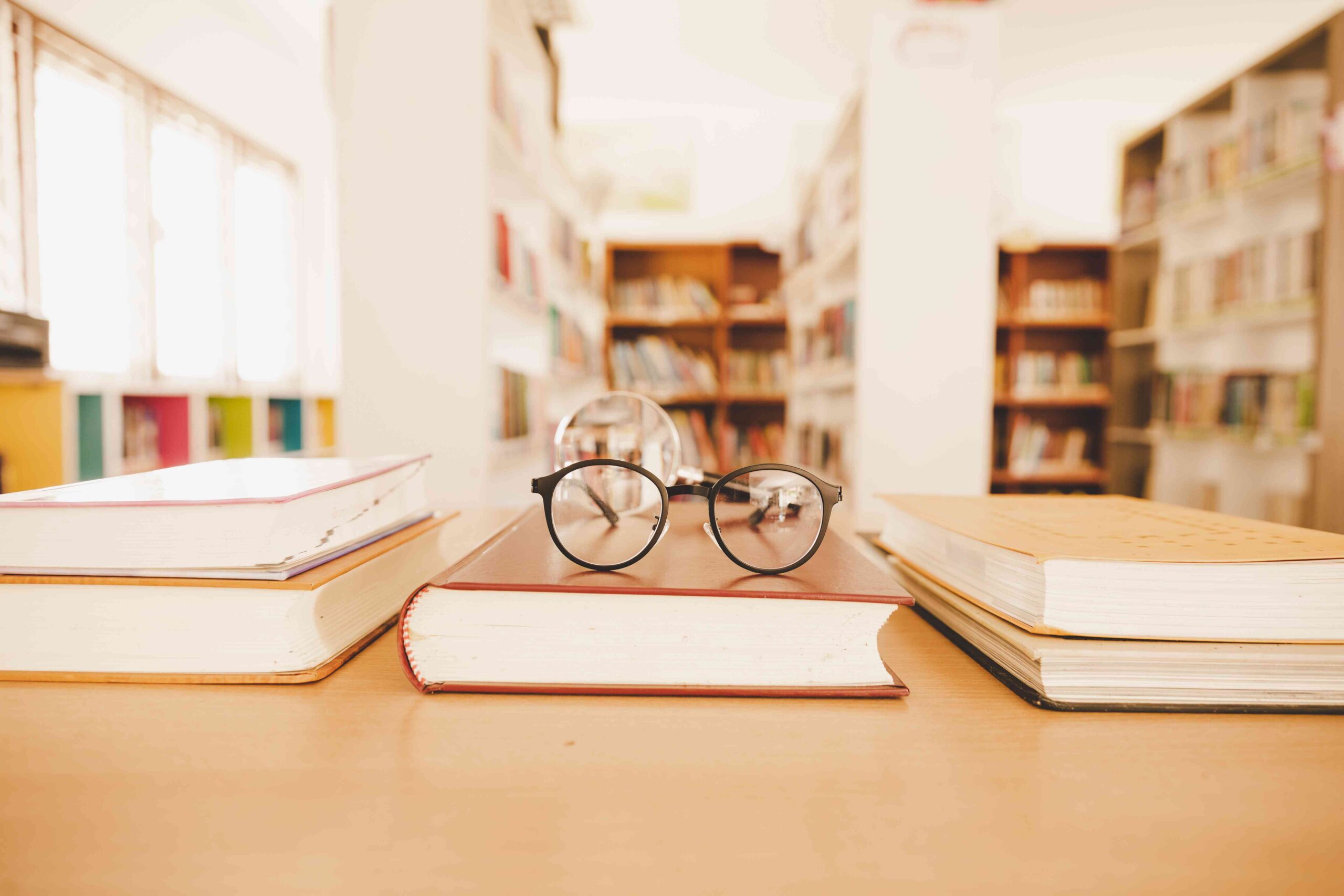 Foto von Büchern auf einem Tisch, daruaf eine Brille. Im Hintergrund Bibliothesräume.